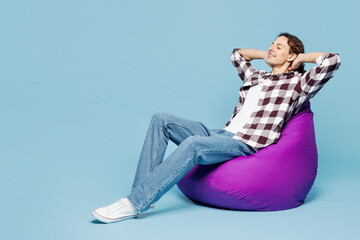 Obraz premium Full body young calm man he wear purple checkered shirt casual clothes sit in bag chair hold hands behind neck isolated on plain pastel light blue cyan background studio portrait. Lifestyle concept.