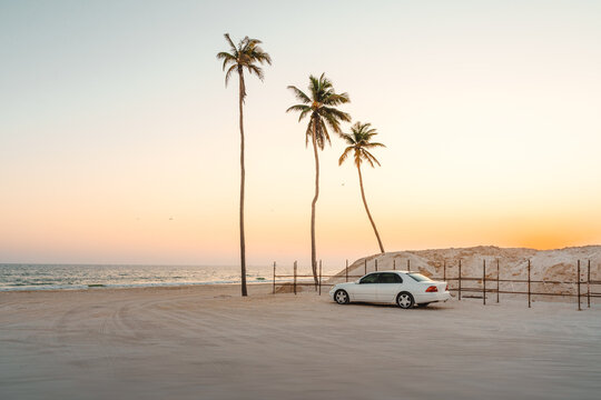 Wei&szlig;es Auto geparkt am Strand mit Palmen bei Sonnenuntergang Salalah Oman