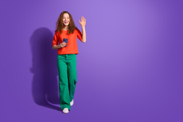 Cheerful young woman in casual outfit waving and holding smartphone against vibrant purple background