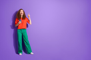 Young woman in colorful fashion with phone, pointing up, on a vibrant purple background, exuding charm and liveliness