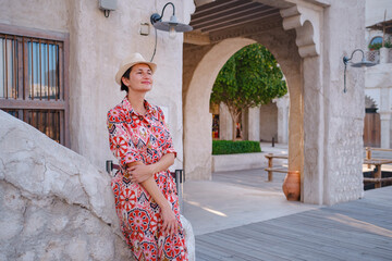 Fototapeta premium Young woman in an ethnic dress and hat exploring Al Seef Park in Dubai. Surrounded by traditional Emirati architecture and modern vibes, she enjoys the charm of Dubai Creek's cultural heritage.