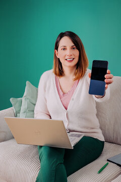 Femme assise sur un canap&eacute; avec ordinateur portable et smartphone, montrant l&rsquo;&eacute;cran du t&eacute;l&eacute;phone.