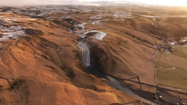 Golden hour aerial view of Sk&oacute;gafoss waterfall in southern Iceland, flowing from the Sk&oacute;g&aacute; River near the base of Eyjafjallaj&ouml;kull volcano, surrounded by rugged, snow-dusted terrain.