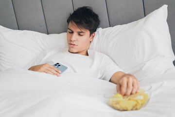Young man relaxing in bed with a smartphone and snacks, enjoying a peaceful and cozy moment in a...
