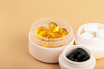 Yellow capsules, green tablets and white pills in open plastic containers on beige background. Concept of health and wellness products or supplement packaging. Vitamins and supplements in open jars