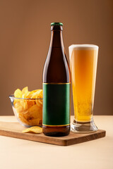 Beer bottle with green label, full beer glass and bowl of potato chips on wooden tray over beige background. Concept of bar ads, party visuals and beer snack combination promotions. Beer with chips
