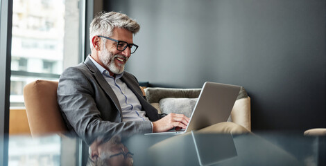 Smiling senior man in suit using laptop, creative photo style, sitting in modern office lounge, concept of remote work and business success. Ai generative