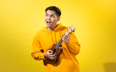 Asian man in yellow hoodie enthusiastically playing a small ukulele against a vivid yellow background. Expressive musical portrait with happy mood and creative, artistic personality