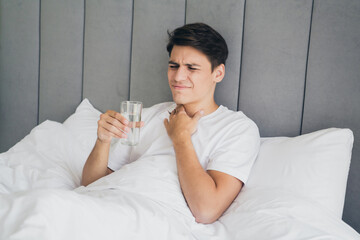 Young man waking up at home holding a glass of water and experiencing a sore throat or discomfort...