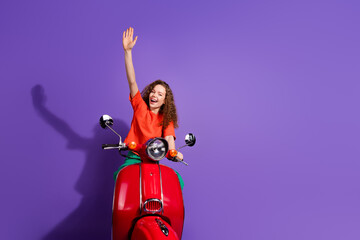 Cheerful young woman on a red scooter waving her hand against a vibrant purple background