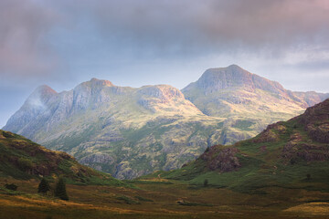 Fototapeta premium A dramatic morning in the Lake District, where golden sunlight bathes the rugged Langdale Pikes, casting a serene glow over the majestic landscape.