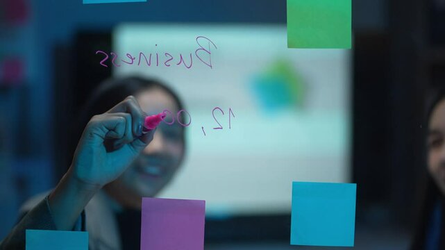 A woman is writing on a white board with a pink marker. The board has a lot of colorful sticky notes on it