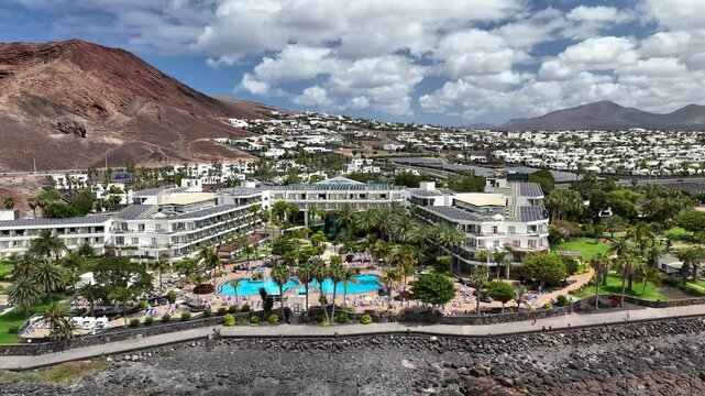Panoramaflug entlang der K&uuml;ste von Playa Blanca, Lanzarote: t&uuml;rkisfarbenes Meer, schwarze Felsen, Hotels, Palmen und vulkanische Landschaften aus der Vogelperspektive.