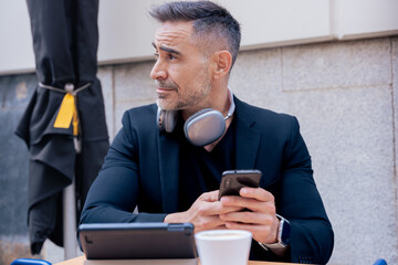 Mature businessman with headphones around neck sits at a cafe table holding a smartphone. Tablet and coffee on the table. He looks aside with a thoughtful expression in a modern urban setting. 