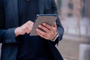 Unrecognizable businessman standing outdoors using a tablet in a modern urban setting. He appears focused and professional. Concept of digital work, technology, and contemporary business lifestyle. 