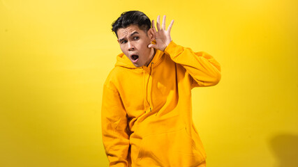 Fototapeta premium Curious young Asian man in yellow hoodie holding his hand to ear as if listening, with surprised expression, against solid yellow background. Studio concept for hearing, attention, or curiosity