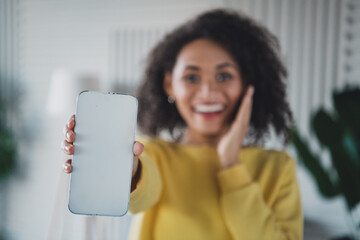 Young woman in yellow pullover holding phone with blank screen and smiling indoors