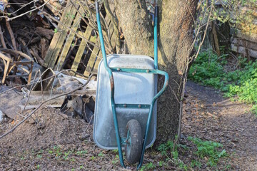 one big empty gray metal wheelbarrow stands near a tree in green grass on the street