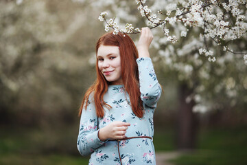Smiling red haired girl sitting on grass in spring park with blooming trees