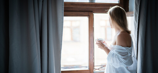 Woman in elegant robe drinking coffee in hotel room and standing near window