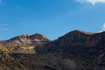 Rugged mountains bathed in warm sunlight, towering over rocky terrain with scattered vegetation, creating a timeless landscape of natural beauty in Fars province.
