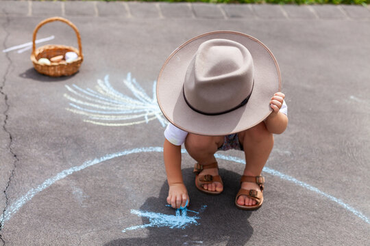 Child drawing with chalk on driveway outside holding onto felt hat