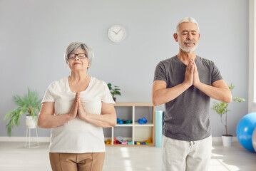 Fototapeta premium Senior people doing yoga. Healthy old couple practicing yoga, meditation and self care together. Relaxed man and woman pensioners in sportswear with closed eyes standing in gym doing namaste gesture