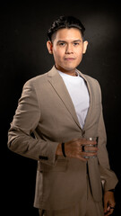 Asian man in beige suit and white t-shirt standing confidently in studio with dark background, holding a clear glass in one hand, ideal for lifestyle, branding, or personal presentation themes