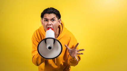 Energetic young Asian man in yellow hoodie shouting through a megaphone with intense expression, standing against a solid yellow background. Concept of announcement, protest, or bold communication
