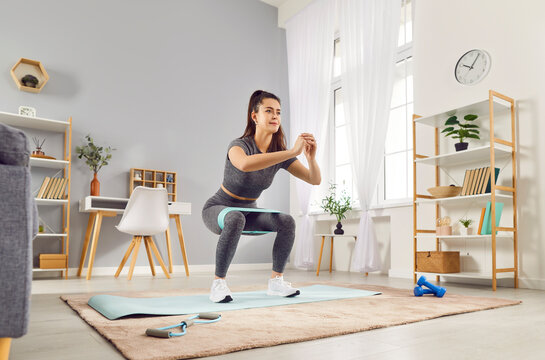 Young sporty woman doing squatting exercises with rubber band standing in living room at home. Happy athletic girl doing domestic workout. Sport, fitness and home training concept.