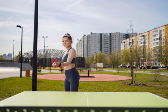A woman is playing table tennis on the street. She's wearing a tracksuit and holding a racket. Residential buildings and trees are visible in the background.