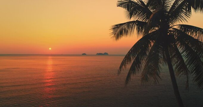 Orange and red sunset over calm sea water with silhouette of palm tree leaves in the foreground and small islands in the distance, Koh Samui, Thailand