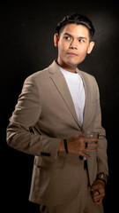 Asian man in beige suit and white t-shirt standing confidently in studio with dark background, holding a clear glass in one hand, ideal for lifestyle, branding, or personal presentation themes