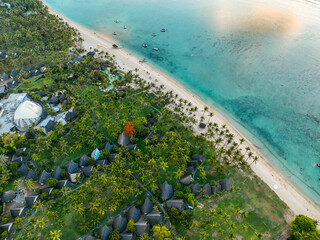 Aerial view of beach resort at sunset with boats, bungalows, and palm trees along the coast