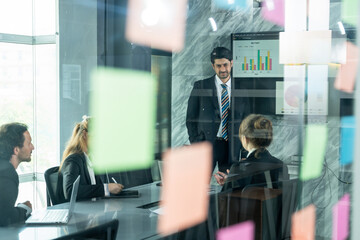 Multicultural business team having conference in modern office behind glass wall sticky notes partially obscuring view presenter explaining chart while team members seated listening and participating