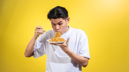 Asian man in a white t-shirt enthusiastically eating noodles from a plate with a fork, looking to...