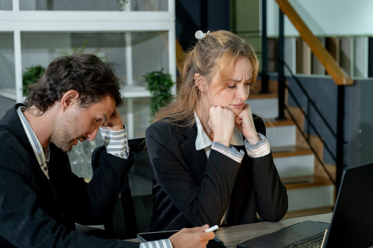 Caucasian man and woman wearing business suits sitting at office desk expressing frustration while looking at laptop screen during business meeting showing stress from corporate work challenges - Powered by Adobe