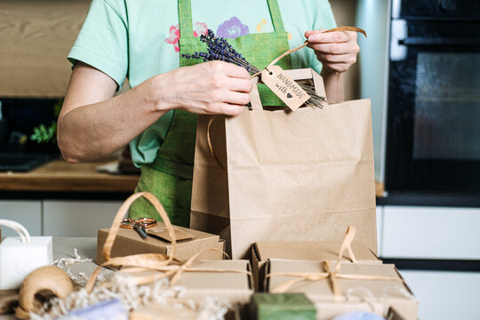 A small business owner ties a Handmade with love tag and lavender sprig to a brown paper bag surrounded by gift boxes. Home-based business, handmade goods