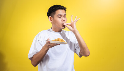 Asian man in a white t-shirt tasting fried noodles with a satisfied expression, eyes closed, and...