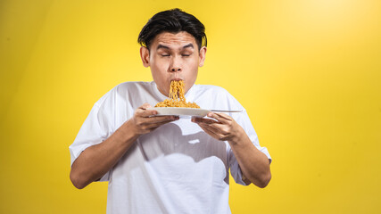 Asian man in a white t-shirt enthusiastically eating noodles from a plate with a fork, looking to...