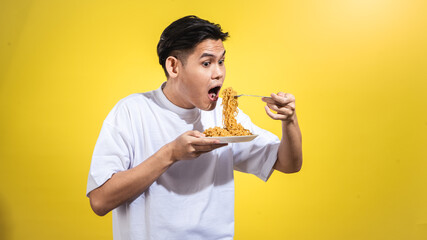 Asian man in a white t-shirt enthusiastically eating noodles from a plate with a fork, looking to...