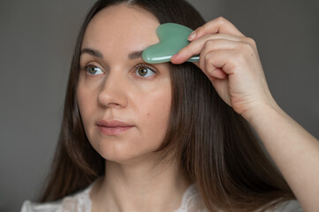 Young woman using a green gua sha scraper for facial massage, improving skin circulation and...
