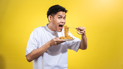 Asian man in a white t-shirt enthusiastically eating noodles from a plate with a fork, looking to...