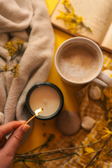 A cozy spring flat lay with a hand lighting a candle, a cup of coffee, a soft blanket, yellow flowers, an open book, and sweet treats on a bright yellow background, creating a peaceful mood