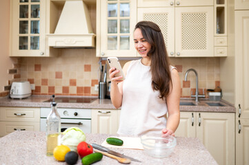 Smiling woman using smartphone while preparing fresh vegetable salad in cozy kitchen. Healthy lifestyle, home cooking and online recipe inspiration