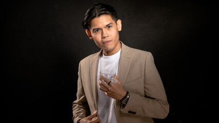 Asian man in beige blazer and white t-shirt holding a transparent glass, looking away with contemplative expression in studio with dark background, perfect for lifestyle or branding visuals