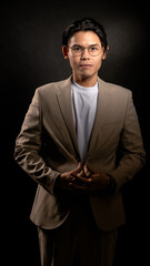 Fototapeta premium Asian man with glasses wearing beige suit and white shirt poses confidently with hands joined in front of chest against dark studio background, ideal for corporate or branding themes