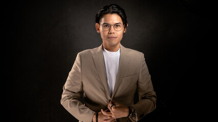 Asian man with glasses wearing beige blazer and white t-shirt standing confidently in studio with dark background, fingers forming a triangle, ideal for personal brand or leadership concept