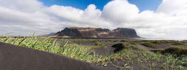Low-angle panorama of windswept black sand dunes in front of rugged Icelandic mountains partially covered by dramatic storm clouds.