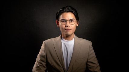Asian man with glasses wearing beige blazer and white t-shirt standing confidently in studio with dark background, fingers forming a triangle, ideal for personal brand or leadership concept